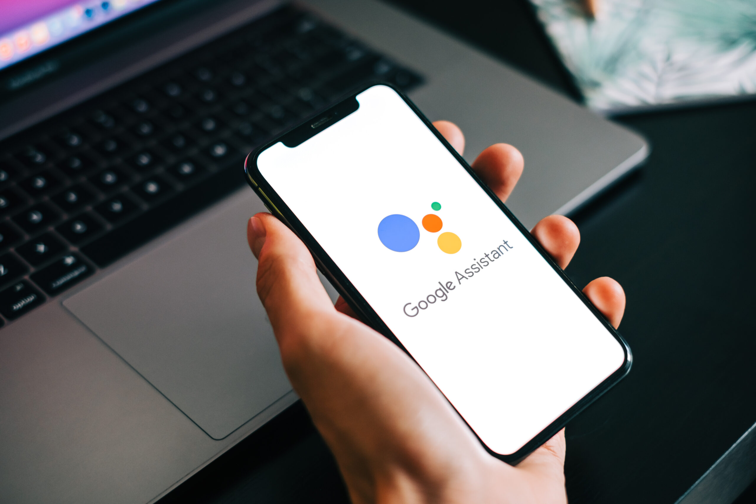 A person holds a phone with the Google Assistant app open in front of their laptop.